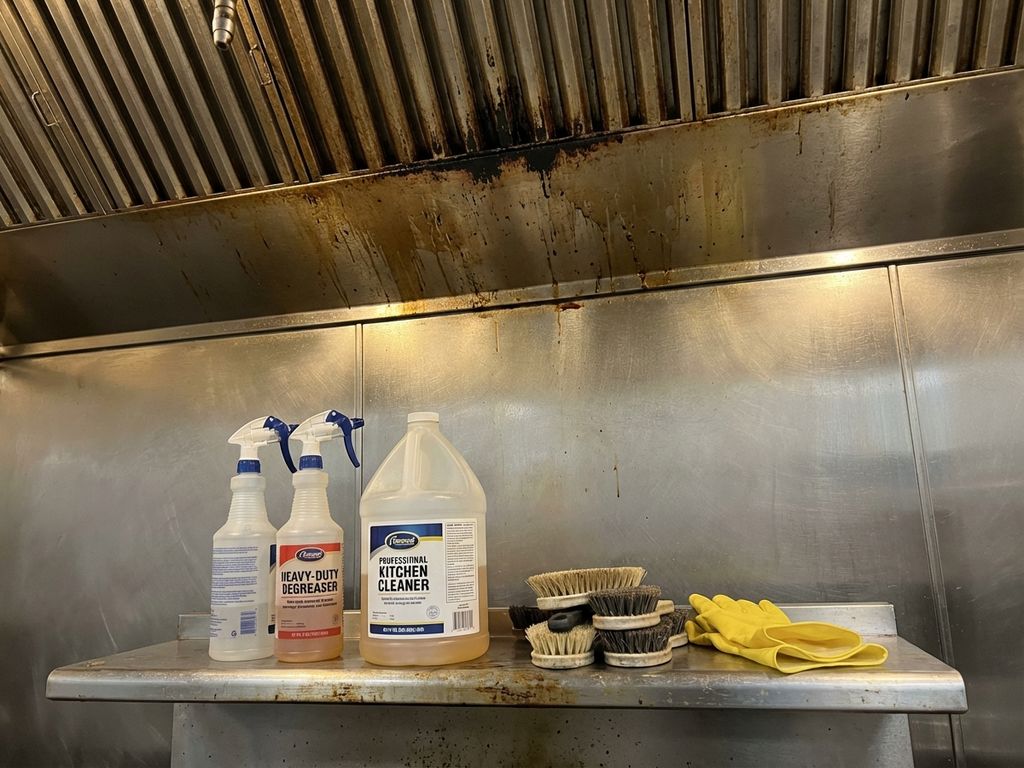 Vettige afzuigkap in restaurantkeuken met schoonmaakspullen en ontvetters op roestvrijstalen aanrecht eronder