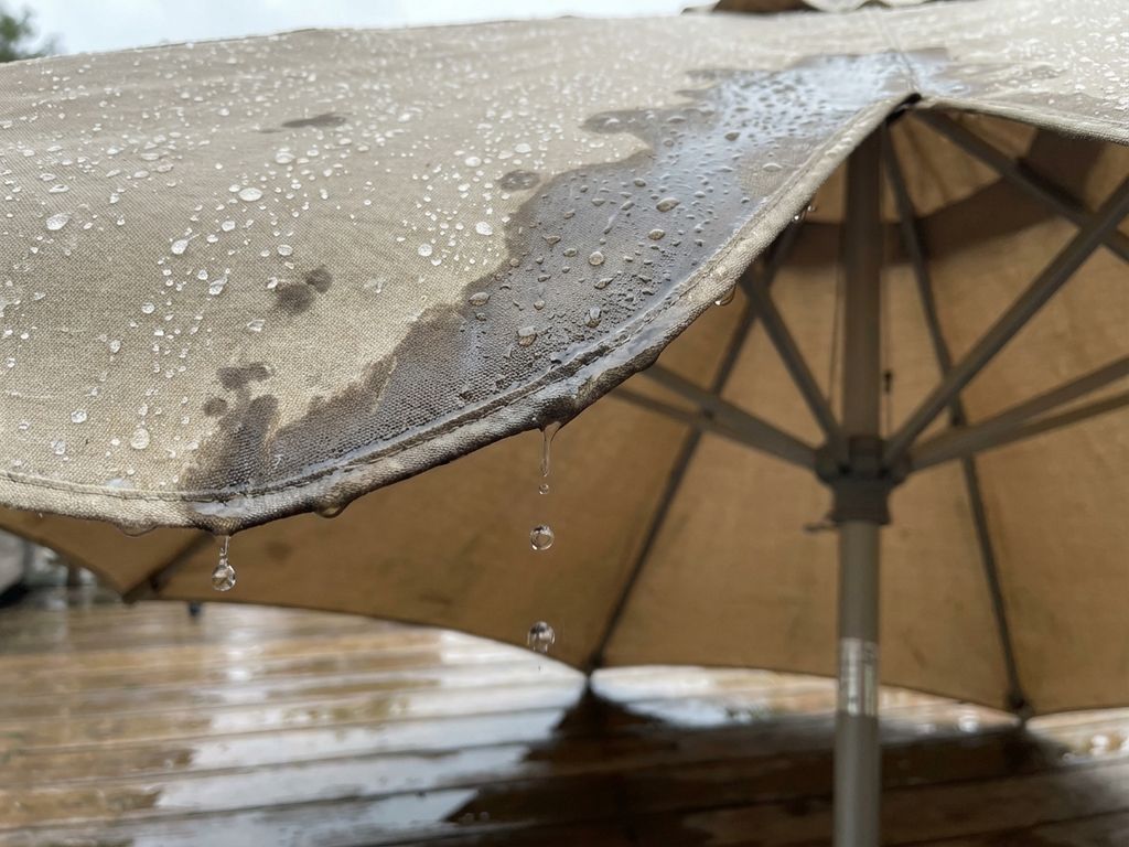 Regendruppels lekken door beige parasoldoek op houten terras, met natte vlekken en plassen zichtbaar
