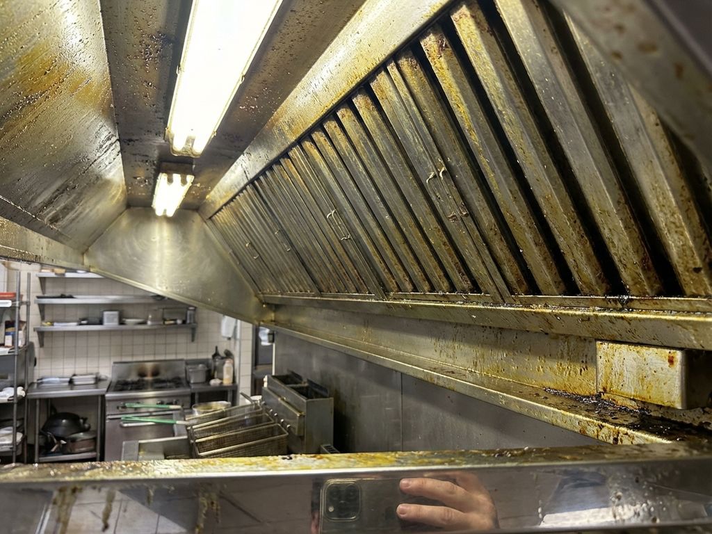 Zwaar vervuilde afzuigkap met geel-bruine vetaanslag in restaurantkeuken, filters en metalen oppervlakken vol vet